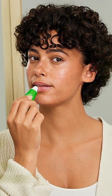 Dark Skin woman holding a Skin Food Lip Balm and applying it to her lips.