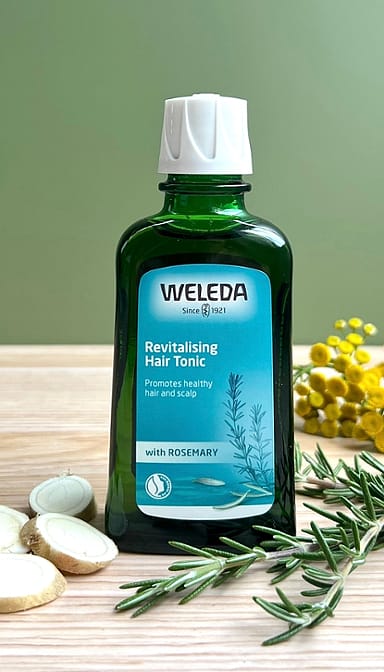 Revitalising Hair Tonic bottle on a sage green background with its ingredinets rosemary leafs and horseradish root.