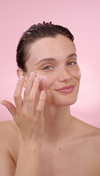 Woman applying Smoothing Wild Rose & White Tea eye cream on her face in a pink background.